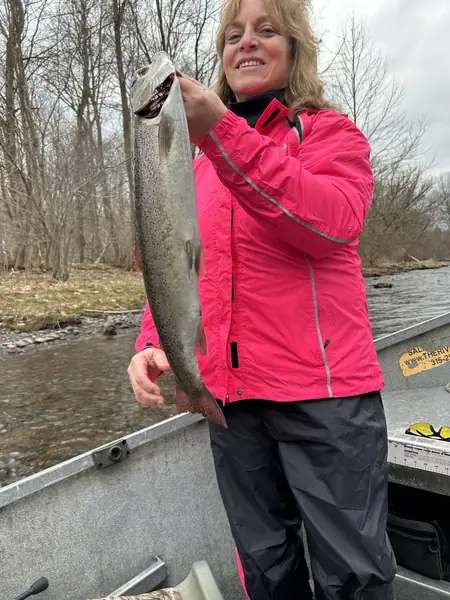 An angler in a pink jacket holds up a silver trout in a drift boat on a river